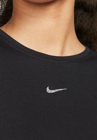 Detailní záběr na krk a horní část hrudi osoby, která má na sobě černé tričko se malým, lesklým stříbrným logem Nike ve tvaru klikatice umístěným přímo pod výstřihem.