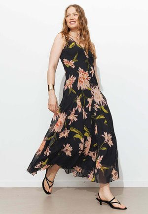 Femme en robe maxi noire à fleurs et sandales noires à brides marchant sur fond blanc, souriante et portant un bracelet manchette doré.