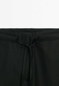 Pantalon en tissu noir montrant la taille avec un cordon noué et des passants de ceinture sur fond blanc.
