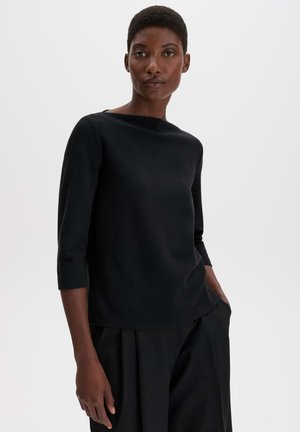Donna con capelli corti che indossa una maglia nera a maniche tre quarti e pantaloni neri, in piedi davanti a uno sfondo chiaro e uniforme.