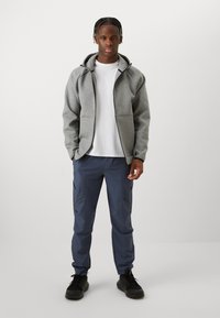 Mann steht und trägt einen grauen Kapuzenpullover mit Reißverschluss, ein weißes T-Shirt, dunkelblaue Hose und schwarze Sneakers vor einem einfarbigen hellen Hintergrund.