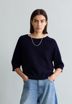 Νέα γυναίκα με κοντά σκούρα μαλλιά φοράει μπλε σκούρο πουλόβερ, ανοιχτόχρωμο τζιν, ασημένιο κολιέ και μικρά σκουλαρίκια κρίκους, με τα χέρια στις τσέπες.