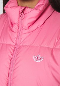 Rosa puffjacka med hög krage, försäkrad med dragkedja och broderad logotyp på bröstet. Slät, glänsande tygstruktur.