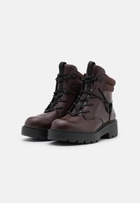 Bottines en cuir marron avec des lacets noirs, col rembourré et semelle épaisse noire. Présente une boucle sur le côté pour un ajustement réglable.