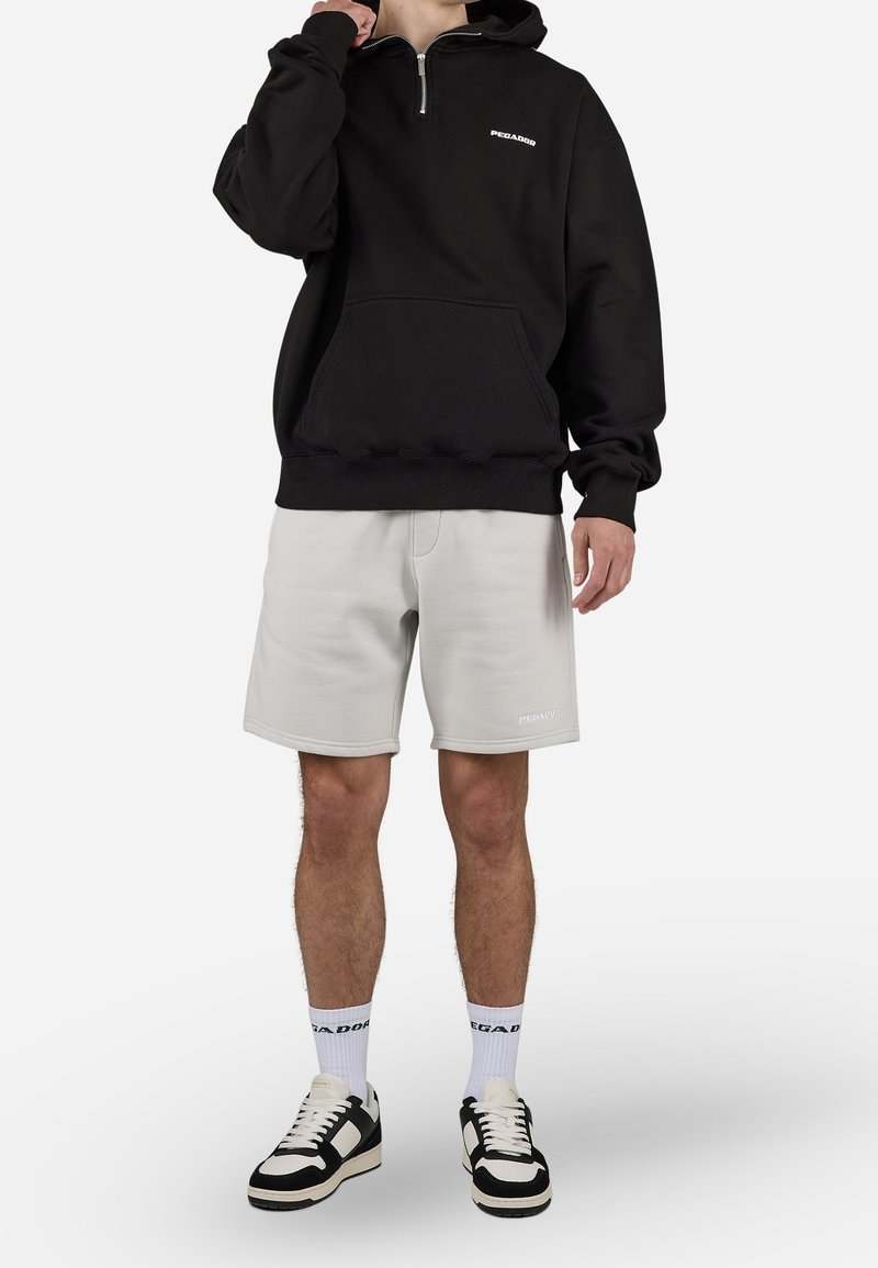 Sudadera con cremallera negra y bolsillo frontal, combinada con pantalones cortos gris claro. Presenta detalles de marca y zapatillas en blanco y negro.