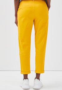 Chinos en coton jaunes avec une coupe droite, ourlets retroussés et deux poches arrière, associés à des baskets blanches sur un fond uni.
