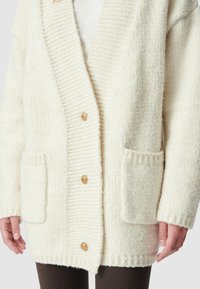 Cardigan lavorato a maglia di colore crema con una texture a costine, dotato di un colletto a scialle, due tasche frontali e tre bottoni dorati decorativi.