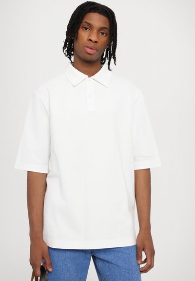 White short-sleeve polo shirt featuring a classic collar and a subtle embroidered detail on the sleeve, worn with blue denim pants.