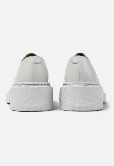 Pari valkoisia korokepohjakenkiä, joissa on kuvioidut paksut pohjat, nähtynä takaa neutraalilla taustalla.