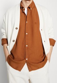 Cardigan crema sopra una camicia button-up color ruggine, con un piccolo accento ricamato. Il tessuto liscio e i bottoni discreti completano il look.