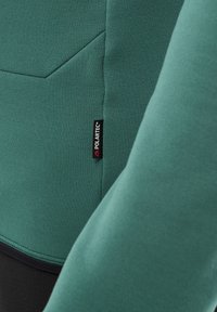 Grüner Fleece-Stoff mit glatter Textur, versehen mit einem sichtbaren schwarzen Polartec-Label. Das Design umfasst minimale Nähte und ein geometrisches Muster.