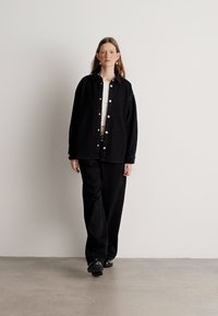Černá oversized košile, jednoduchý bílý top, černé volné kalhoty, v kombinaci s černými botami. Hladká textura s detaily stříbrných knoflíků. Neutrální pozadí.