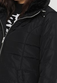 Svart pufferjacka med dragkedja, med en plyschfodrad svart huva, quiltad textur och snedställda sidofickor.