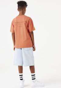Camiseta de manga corta color óxido con texto impreso, combinada con pantalones cortos de mezclilla azul claro y zapatillas blancas, acentuada con calcetines a rayas en blanco y negro.