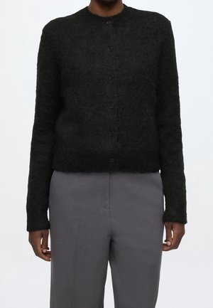 Personne portant un cardigan noir duveteux à boutons avec manches longues et un pantalon taille haute gris sur un fond uni.