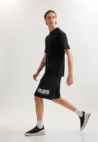 Camiseta negra y pantalones cortos negros con gráfico de "ILIMITADO", usados con zapatillas deportivas negras y calcetines tobilleros blancos; textura suave, diseño casual.