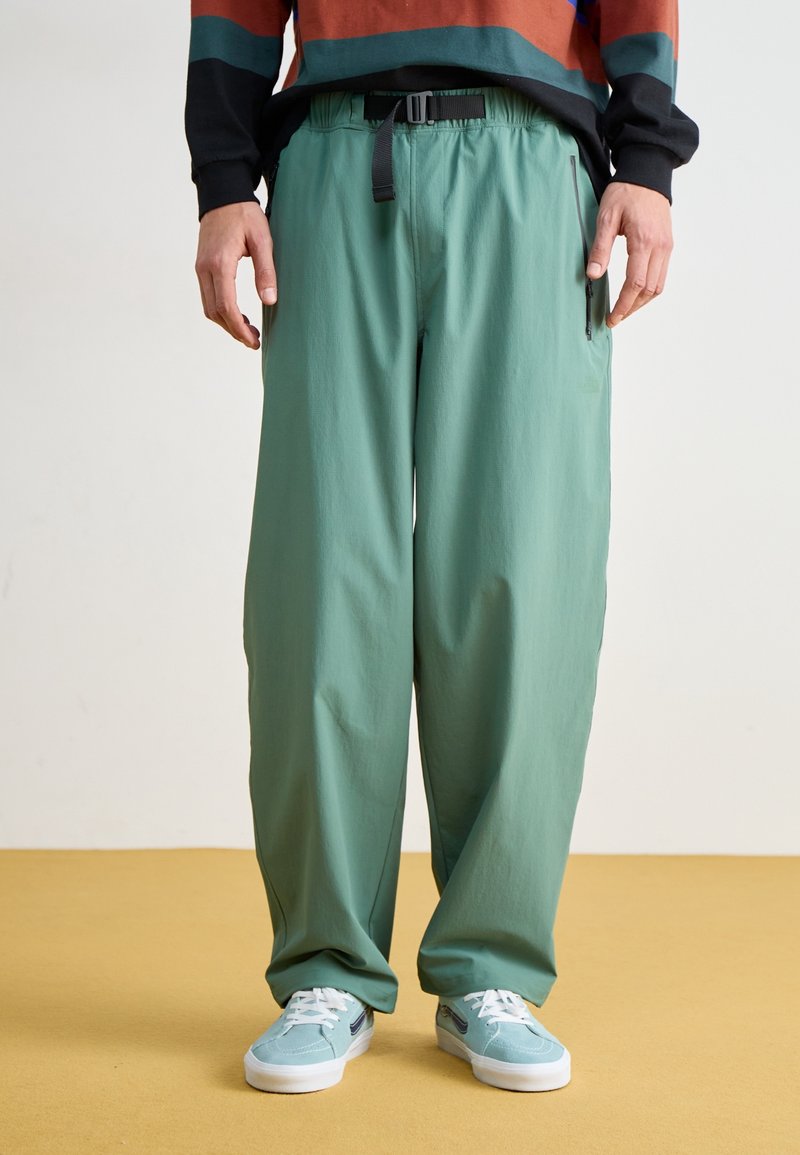 Personne portant un pantalon ample vert clair avec une ceinture noire réglable et des baskets bleu clair, debout sur un sol jaune.