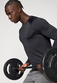 Μαύρο αθλητικό μπλουζάκι με μακριά μανίκια και στρογγυλή λαιμόκοψη, συνδυασμένο με γκρι παντελόνι γυμναστικής, κρατώντας μπάρα με μαύρες δίσκους βάρους.