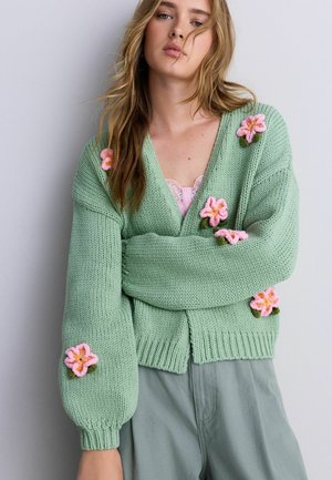 Frau trägt eine grüne, gestrickte Strickjacke, verziert mit pinken gehäkelten Blumen, kombiniert mit grauer Hose und einem pinken Spitzen-Top darunter.