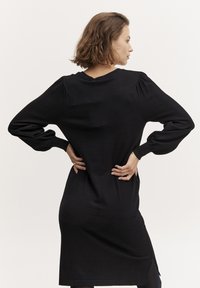Abito nero in maglia con maniche lunghe e puffate e scollatura a V. Presenta una silhouette aderente e un orlo all'altezza del ginocchio con spacchi laterali.