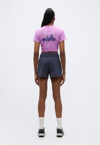 Γυναίκα που στέκεται με την πλάτη γυρισμένη, φορώντας μοβ μπλούζα Patagonia με γραφικό βουνών, σκούρα μπλε σορτς, λευκές κάλτσες και μαύρα αθλητικά παπούτσια σε λευκό φόντο.