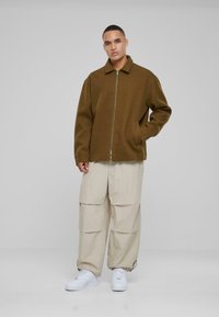 Brown zip-up jacket with a collar, worn over beige cargo pants. The outfit features a relaxed fit and white sneakers. Simple, functional design.