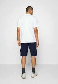 T-shirt en coton blanc à manches courtes et col rond, associé à un short cargo bleu marine. Le modèle porte des chaussettes grises et des baskets blanches.
