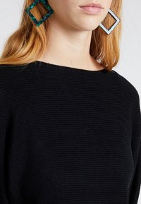 Zwarte gebreide trui met een brede halslijn; model draagt groene en blauwe geometrische oorbellen met vierkante vormen. Glad textuur zichtbaar.