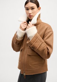 Bruine wolmix jas met een hoge kraag, zachte witte fleece voering, twee voorzakken en ritssluiting. Accentueert met knoopdetails.