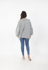 Grijze oversized trui met verlaagde schouders, ribbelhalslijn en een relaxte pasvorm, gecombineerd met blauwe skinny jeans en doorzichtige sandalen met hak.