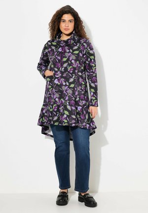 Tunique longue florale avec un col drapé, ornée de motifs violets et verts sur fond noir, assortie à un jean et des chaussures noires.