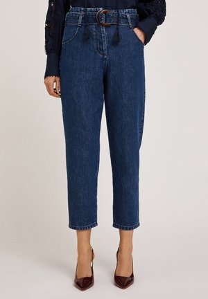 Woman wearing high-waist blue denim jeans with a brown belt and maroon pointed-toe heels, hands in pockets, beige background.