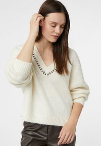 Kremowy sweter z dekoltem w serek i luźnymi rękawami, z teksturowanym wzorem dzianiny. Dolna krawędź jest ściągana na drutach, z subtelnym dekoracyjnym ściegiem wokół dekoltu.