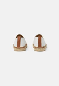 Par de espadrilles blancas con lengüetas de cuero marrón en el talón, plantillas de yute trenzado y suelas de goma, mostradas desde atrás sobre un fondo blanco.