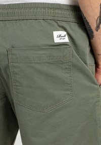 Bermudas verde oliva hechas de una mezcla de algodón, con un bolsillo trasero con costuras reforzadas y una etiqueta blanca con la marca "Reell".