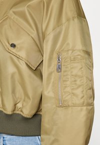 Jachetă bomber khaki cu tiv verde cu nervuri, buzunar cu fermoar pe mânecă cu accesorii argintii și țesătură netedă, texturată. Ajustare personalizabilă.
