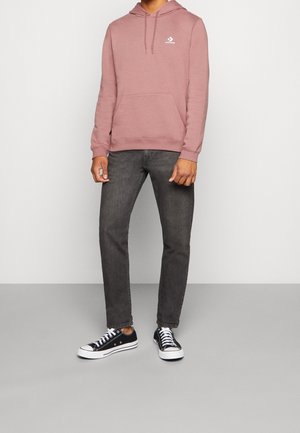 Persona con sudadera con capucha Converse color rosa polvo, jeans gris oscuro y zapatillas Converse negras de caña baja, de pie sobre un suelo gris frente a una pared blanca.