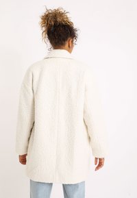 Krämfärgad kappa i ett texturerat fleece-material, avslappnad passform, stor krage och långa ärmar, stylad med ljusblå jeans.