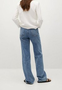 Jean bleu en denim taille haute avec une coupe droite, doté de poches arrière et d'une texture délavée. Porté avec une chemise blanche et des sandales.