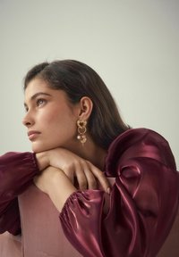 Goldene Ohrringe mit herzförmigen Akzenten, bunten Perlen und einem Perlenanhänger. Das Model trägt ein kastanienbraunes Satinoberteil mit voluminösen Ärmeln.