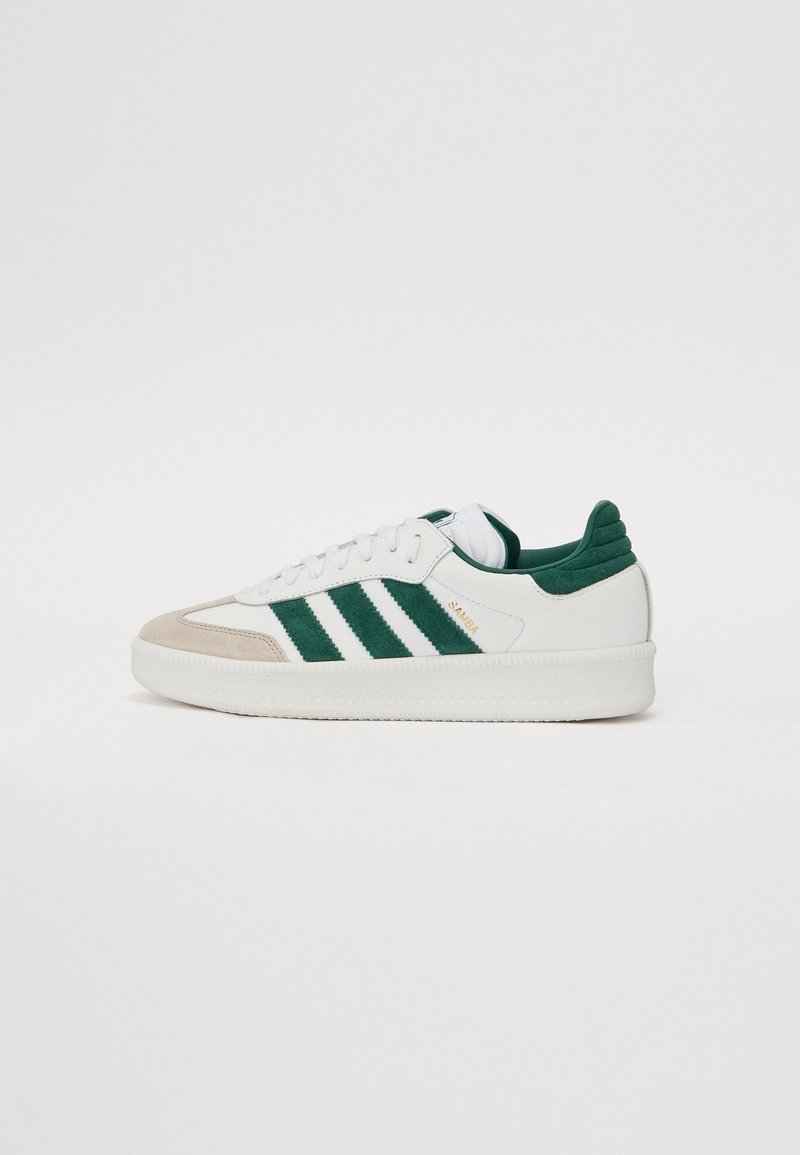 Hvit sneaker med grønne og beige aksenter, med tre grønne striper, strukturert semsket tå og hæl, og en glatt gummisåle.