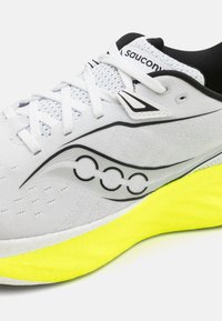 Weißer Sportschuh mit Mesh-Obermaterial, schwarzen Akzenten und reflektierendem Logo. Verfügt über eine leuchtend gelbe Sohle für Kontrast und Dämpfung.