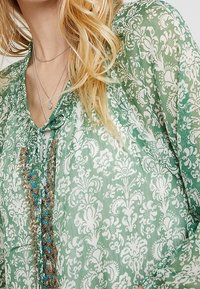 Grönmönstrad blus i lättviktsmaterial med blommönster, dragsko i halsringningen och pärlaccenter längs framsidan.