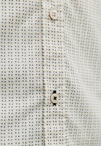 Chemise blanche avec un petit motif à pois noirs et beiges, comportant deux boutons ronds beiges sur la patte de boutonnage avec une couture en fil noir.