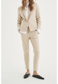 Saco y pantalones de traje beige en tejido suave, combinados con una camisa blanca. Zapatos negros tipo mocasín con un acabado texturizado y detalle de cordones.