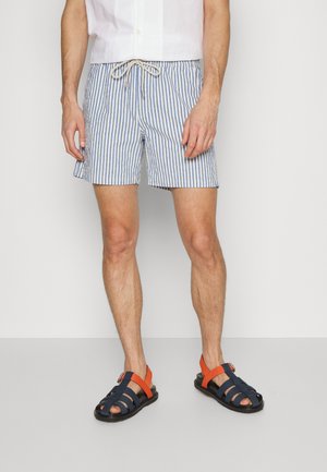 Striped swim shorts in blue and white cotton with an elastic waistband and drawstring. Paired with navy sandals featuring orange accents.