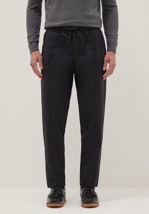 Hombre con pantalones negros con cordón combinados con zapatos negros de cordones y una camiseta de manga larga gris, de pie frente a un fondo beige.