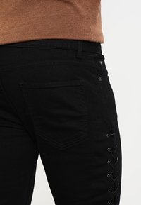 Jeans de mezclilla negra con un bolsillo en la parte posterior y detalles de cordones a lo largo del lado. El material es elástico, con una textura suave y un diseño ajustado.