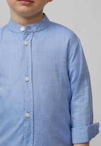 Lichtblauw overhemd met een Mao-kraag, met lange mouwen die zijn opgerold en witte knopen aan de voorkant.