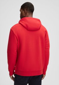 Felpa rossa con una texture morbida, cappuccio regolabile e polsini a costine. Presenta una vestibilità classica con schiena liscia e senza loghi visibili.